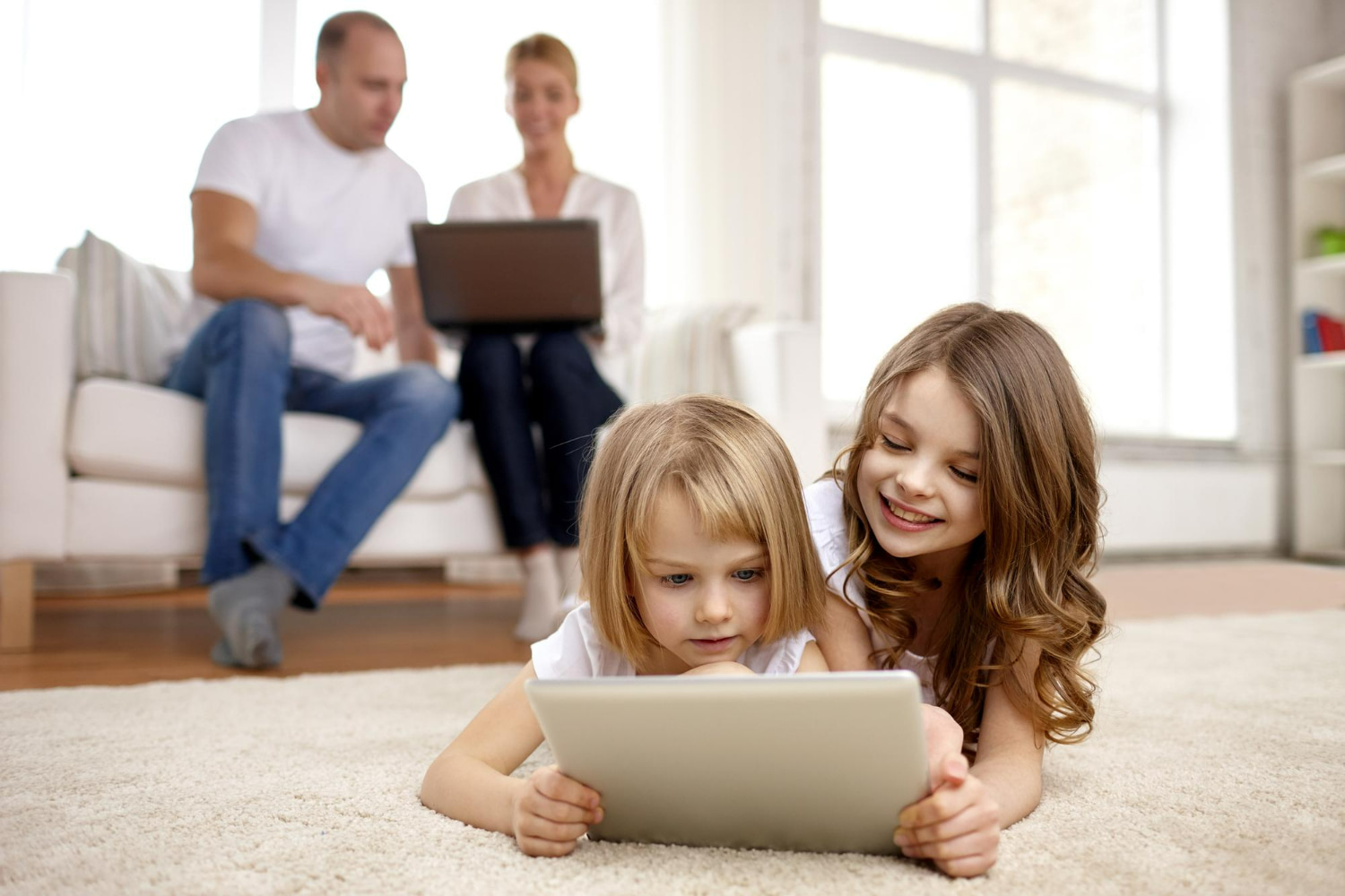 Two sisters lie together on a cozy carpet in a bright, modern living room, smiling and looking at a tablet screen. Two sisters lie together on a cozy carpet in a bright, modern living room, smiling and looking at a tablet screen.