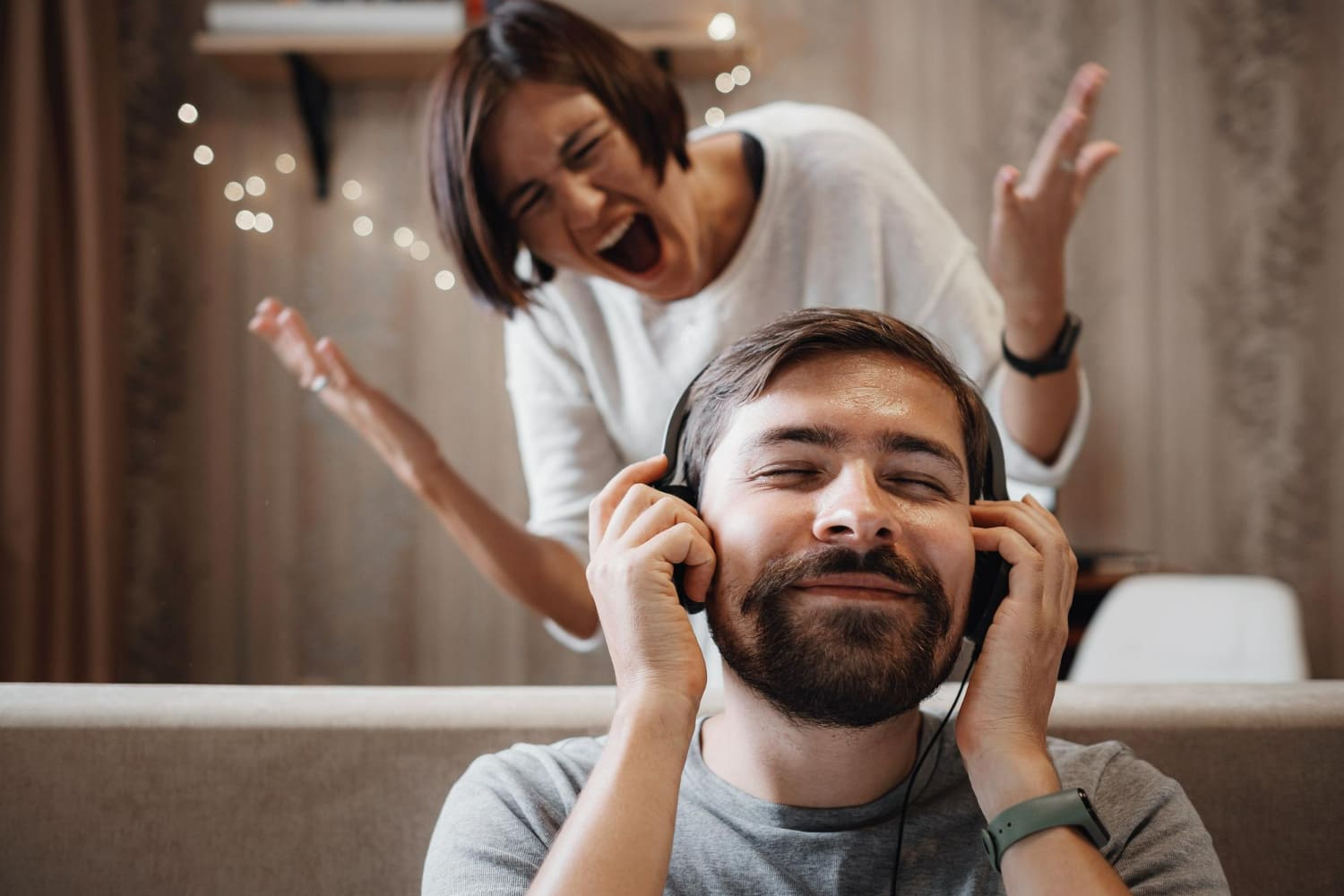 A smiling man wearing headphones while a frustrated woman shouts behind him, illustrating emotional disconnection, relationship conflict, avoidance, poor communication, and ignoring a heated argument. A smiling man wearing headphones while a frustrated woman shouts behind him, illustrating emotional disconnection, relationship conflict, avoidance, poor communication, and ignoring a heated argument.