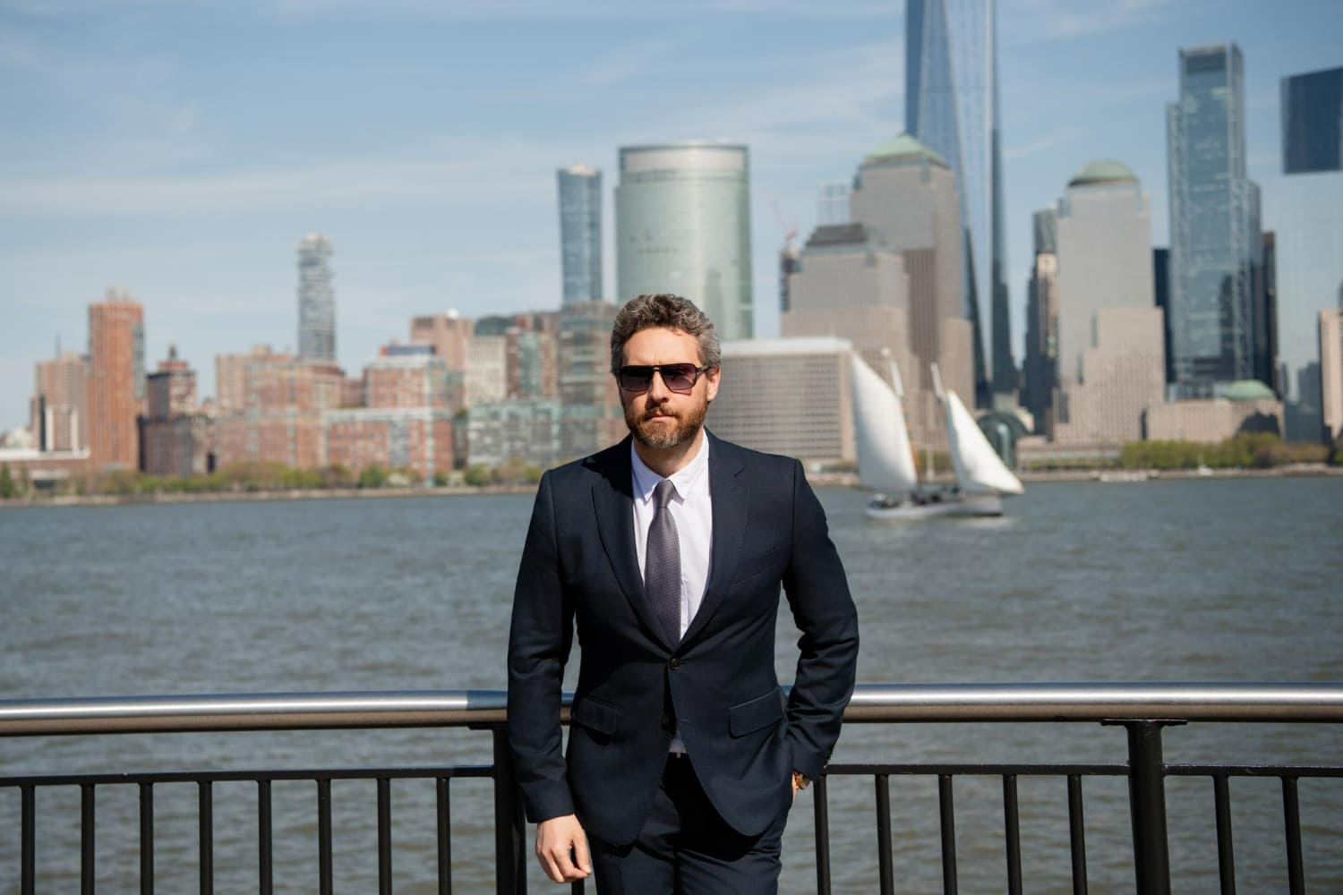 A well-dressed man with gray, curly hair and a full beard stands confidently by a waterfront railing, wearing a dark navy suit, white dress shirt, and gray patterned tie, with sunglasses adding a sharp, composed edge to his expression. A well-dressed man with gray, curly hair and a full beard stands confidently by a waterfront railing, wearing a dark navy suit, white dress shirt, and gray patterned tie, with sunglasses adding a sharp, composed edge to his expression.