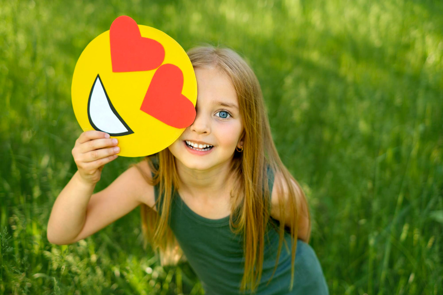 A cheerful young girl with long hair peeks out from behind a handmade paper cutout of a heart-eyes emoji, covering one eye while flashing a big genuine smile.