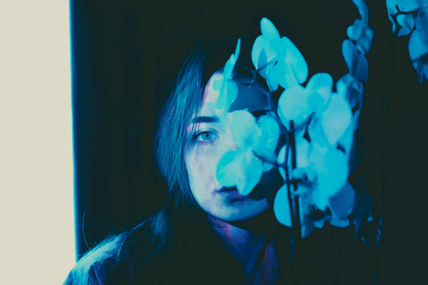 A moody, cyanotype-toned portrait of a young woman with long dark hair, partially obscured by a branch of pale orchid blossoms in the foreground.