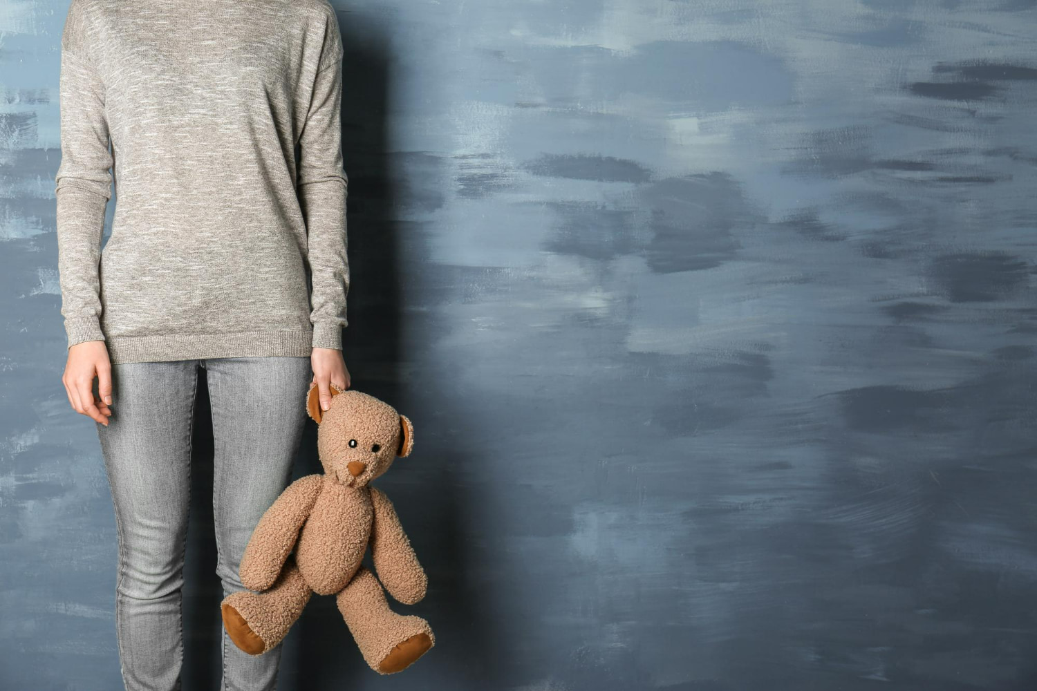 A cropped, faceless shot of a person standing against a textured blue-gray wall, dressed casually in a heather sweater and jeans, holding a teddy bear loosely by one paw at their side.