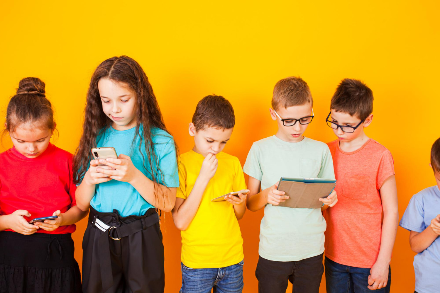 A row of children in colorful t-shirts stand side by side against a bright orange background, each completely engrossed in their own device A row of children in colorful t-shirts stand side by side against a bright orange background, each completely engrossed in their own device