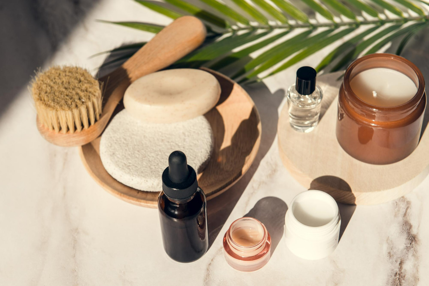 An overhead flat lay of natural self-care essentials — a wooden body brush, pumice stone, bar soap, dropper serum bottle, open cream jars, a glass perfume bottle An overhead flat lay of natural self-care essentials — a wooden body brush, pumice stone, bar soap, dropper serum bottle, open cream jars, a glass perfume bottle