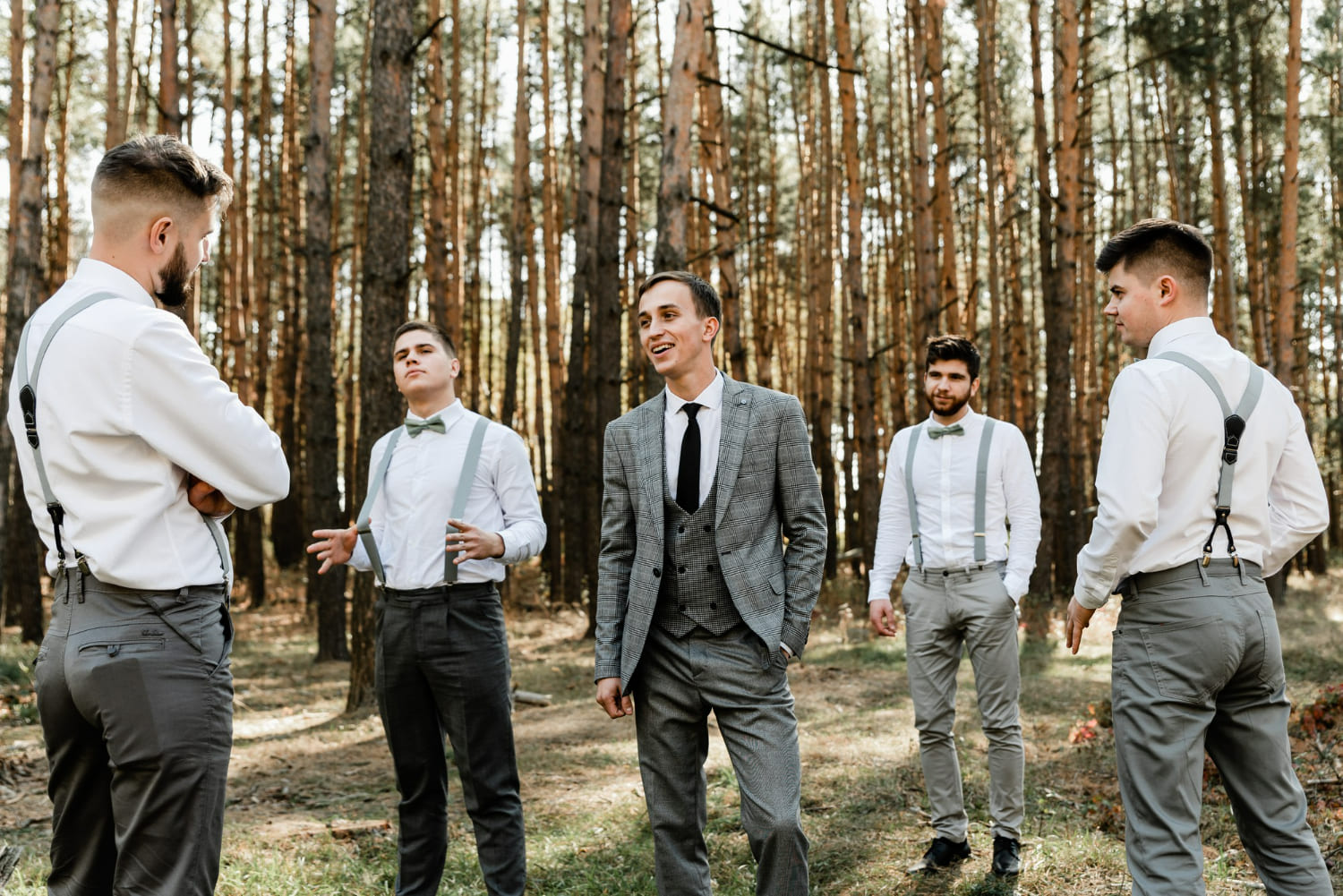 A groom in a gray plaid suit stands centered among four groomsmen dressed in white shirts, suspenders, and bow ties, gathered together in a tall pine forest bathed in soft natural light.