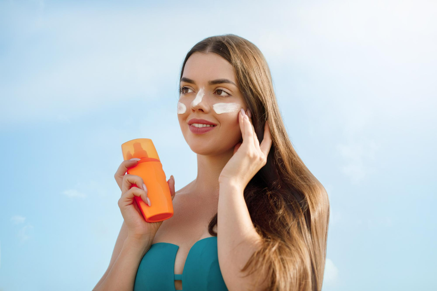 A smiling woman in a teal swimsuit holds an orange sunscreen bottle while applying white sun protection cream to her cheek and nose, set against a bright blue sky background. A smiling woman in a teal swimsuit holds an orange sunscreen bottle while applying white sun protection cream to her cheek and nose, set against a bright blue sky background.