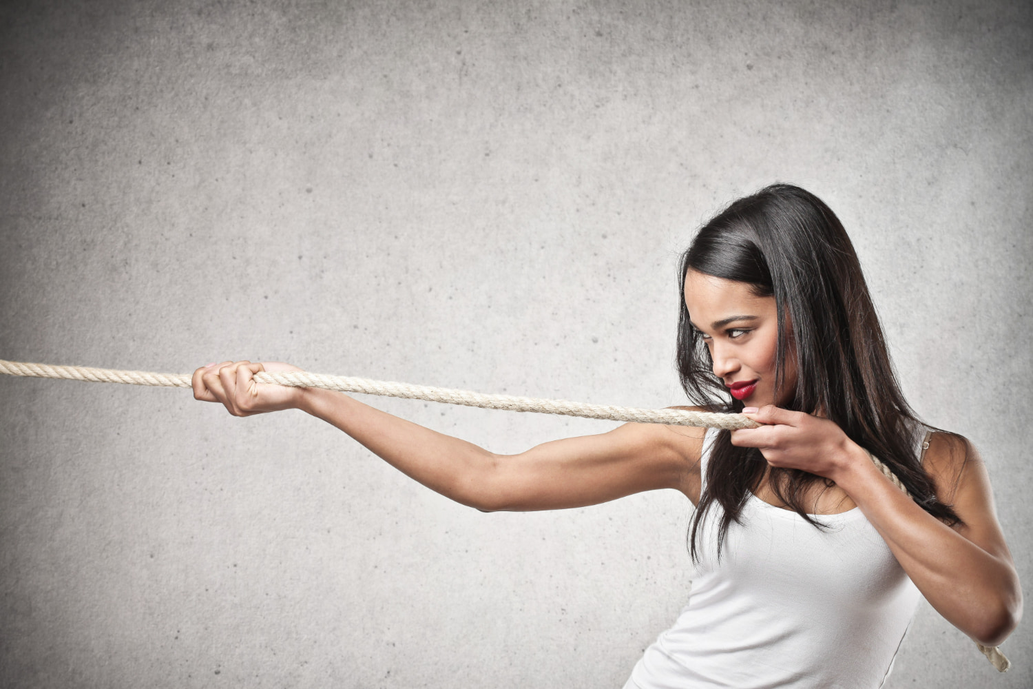 A studio portrait of a young woman with long dark hair and red lipstick, wearing a white tank top, pulling hard on a thick rope against a plain grey textured backdrop. 