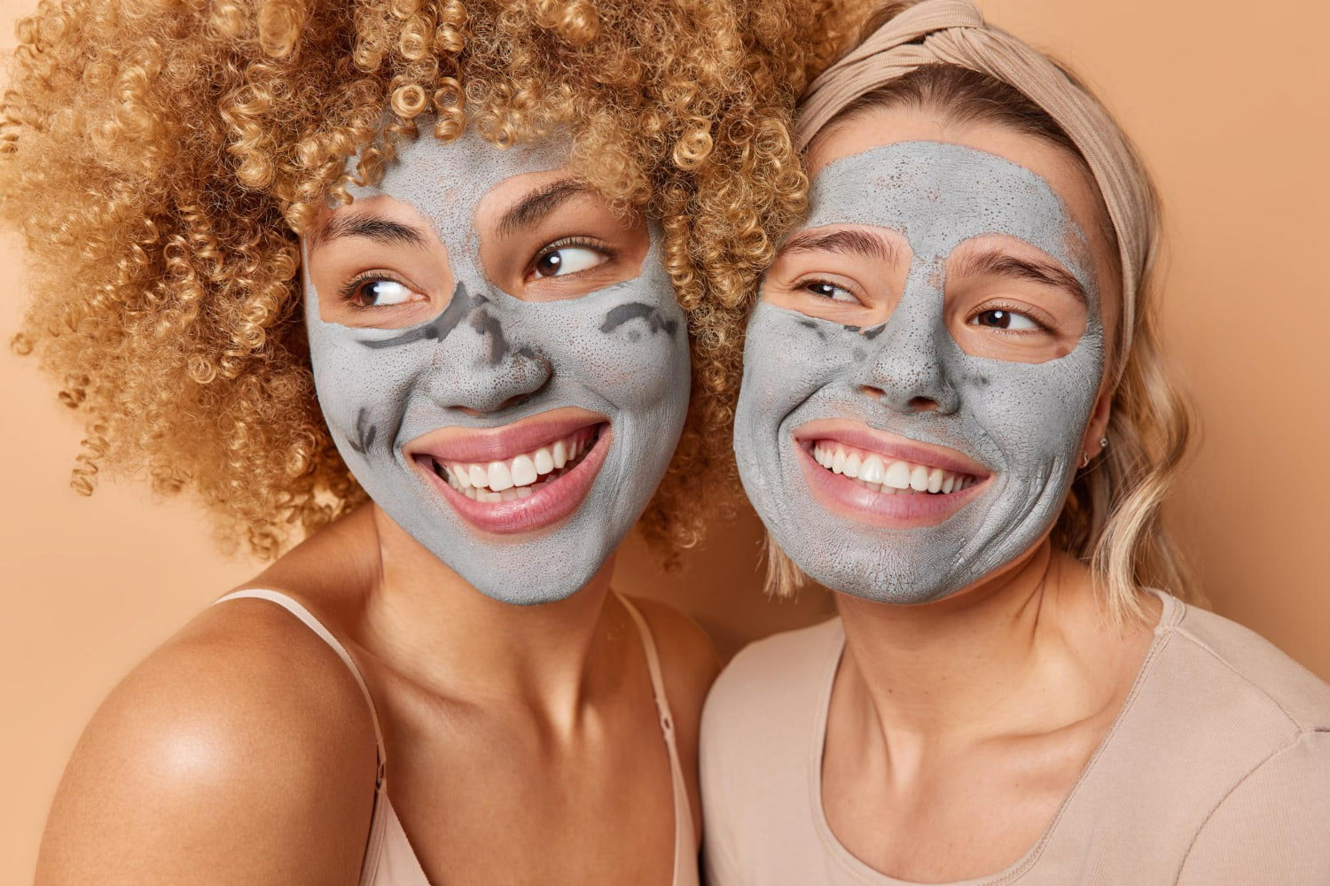 Two women with clay face masks applied smile widely while glancing playfully to the side, posed close together against a warm neutral background.