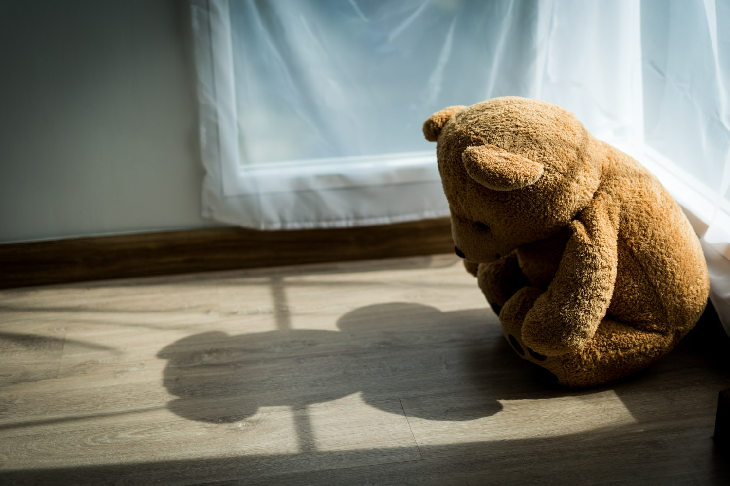 A tan teddy bear sits alone on a wooden floor beside a sunlit window, its head slumped forward in a pose suggesting sadness or abandonment.