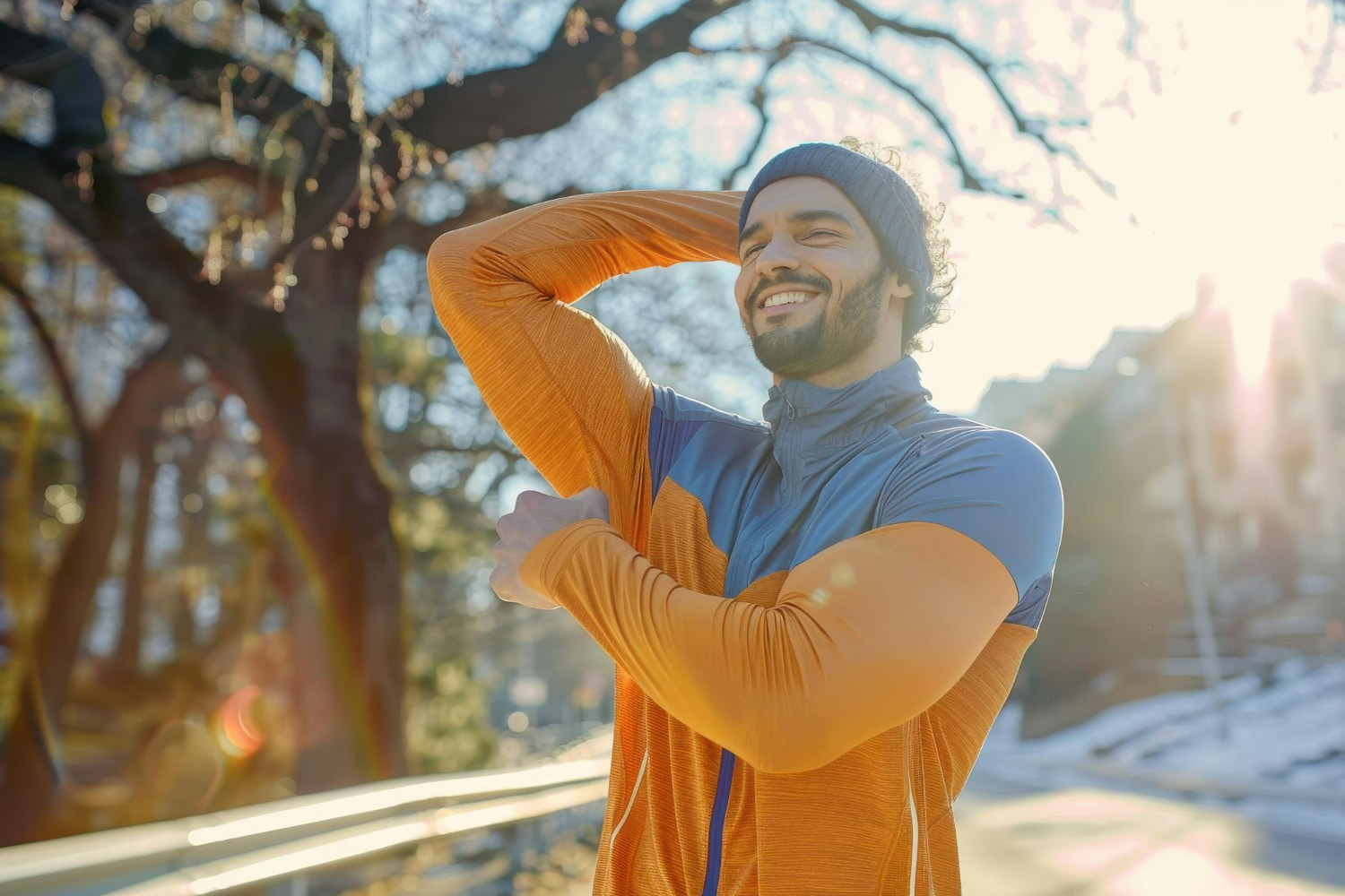 A smiling bearded man in an orange athletic jacket and beanie stretches his arms outdoors on a sunny winter morning, surrounded by bare trees and soft light.