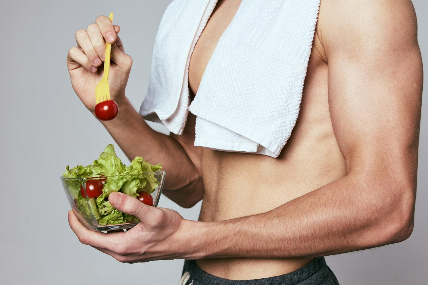 A muscular man with a towel around his neck eats a fresh salad with cherry tomatoes and lettuce straight from the bowl after a workout.