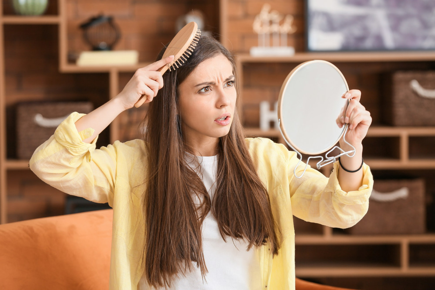 A young woman with long, dark brown hair examines her strands with concern, holding a hand mirror while brushing through her hair.