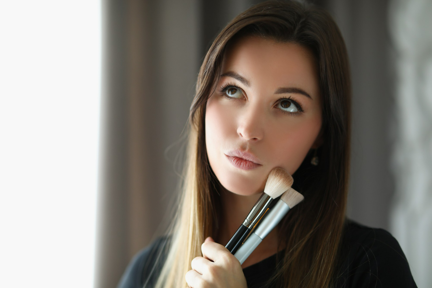 A portrait of a contemplative woman with long brunette hair holding professional makeup brushes, captured in soft natural light.