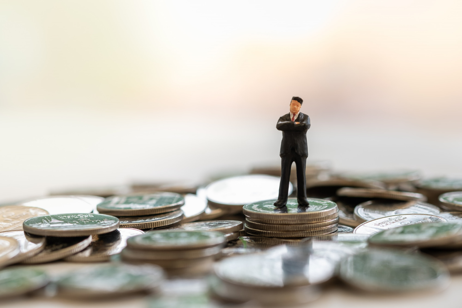 A miniature businessman in formal business attire stands confidently atop a towering stack of coins, arms crossed in a posture of authority and achievement.