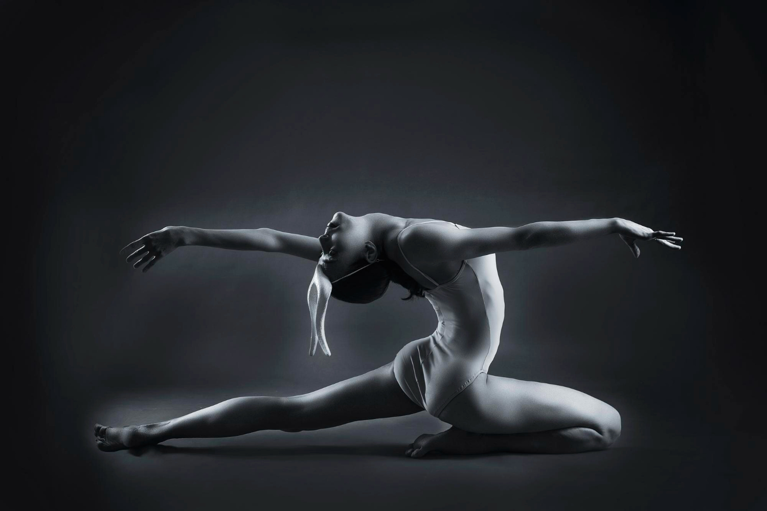 A striking monochrome studio photograph captures a dancer in a deep backbend split pose, arms extended wide with head thrown back, sculpted against a dark, moody background.