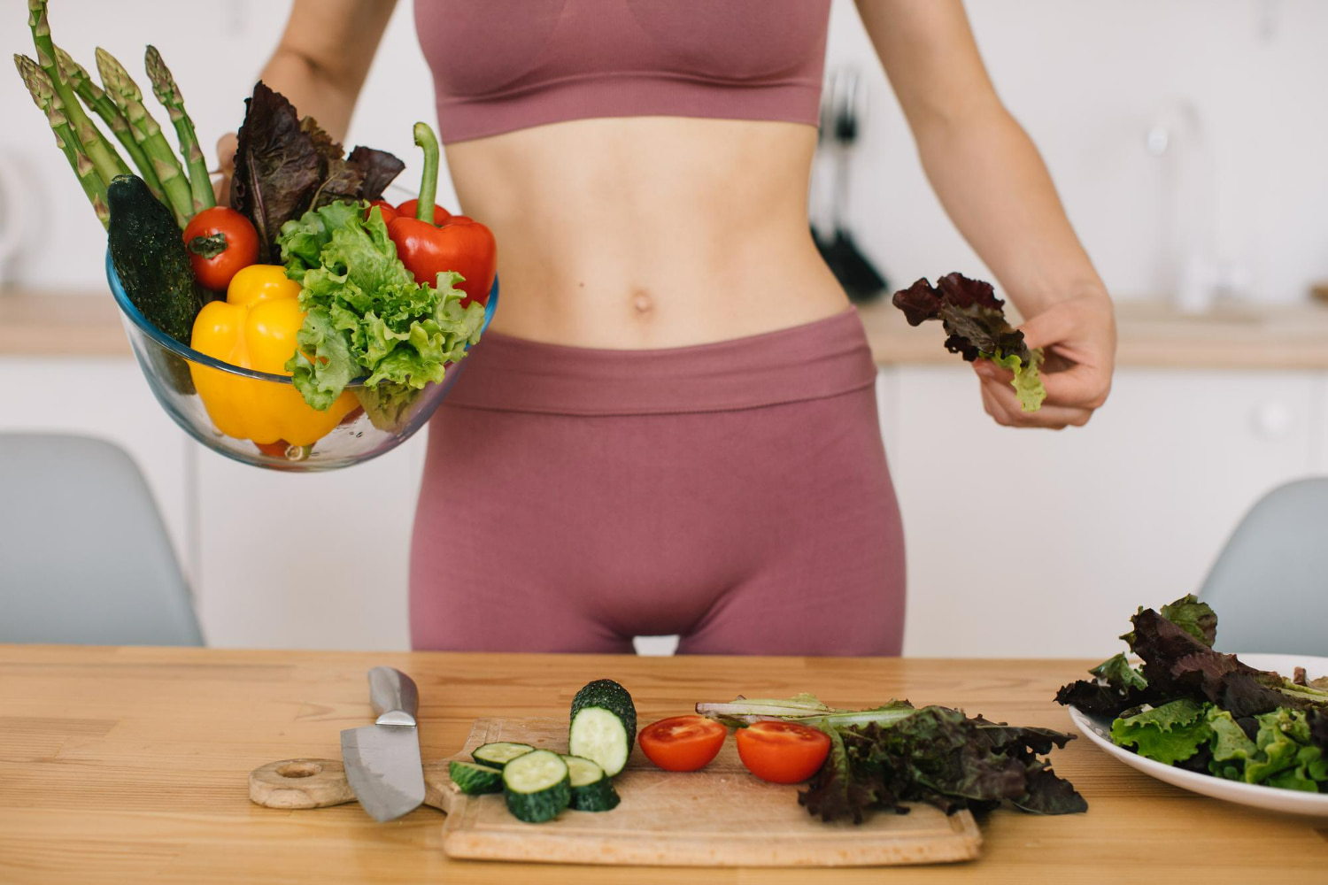 A woman in athletic wear holds a bowl of fresh vegetables while preparing a healthy salad at a wooden kitchen table, surrounded by cucumbers, tomatoes, peppers, and greens.