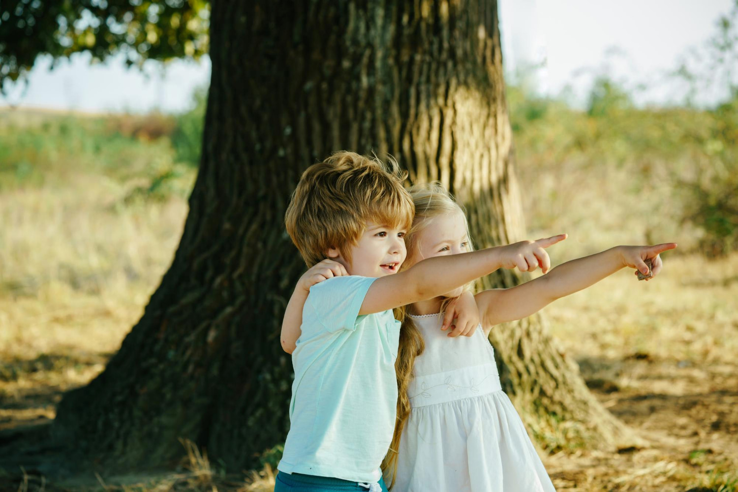 This charming outdoor portrait captures two young children in a moment of pure joy and curiosity, standing beneath the shelter of a magnificent old tree.